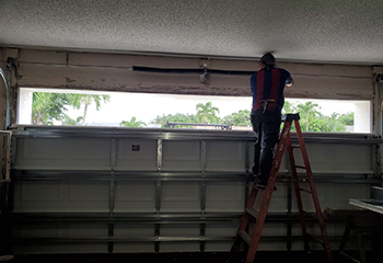 2. Technician on a ladder replacing a garage door spring above a partially open white sectional door, with palm trees visible outside in West Valley City.