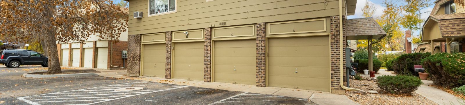 A row of beige residential garage doors with brick pillars in Magna, surrounded by autumn trees and a quiet neighborhood street.
